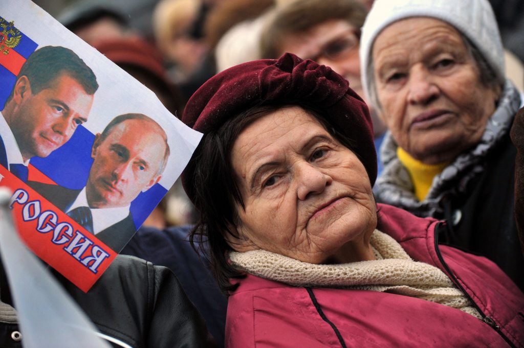 An elderly woman stands next to a flag depicting Russian President Vladimir