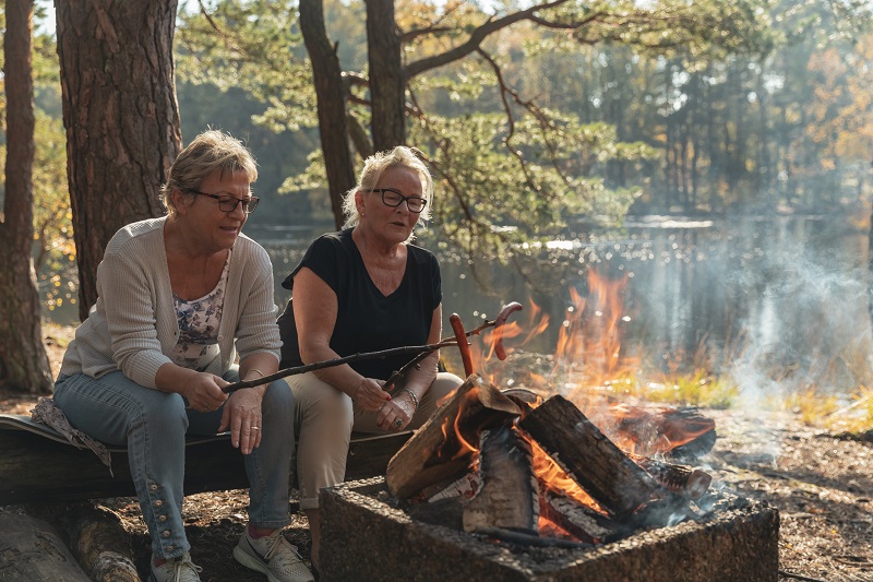 To godt voksne kvinner griller pølser på et bål i skogen, rett ved et vann.