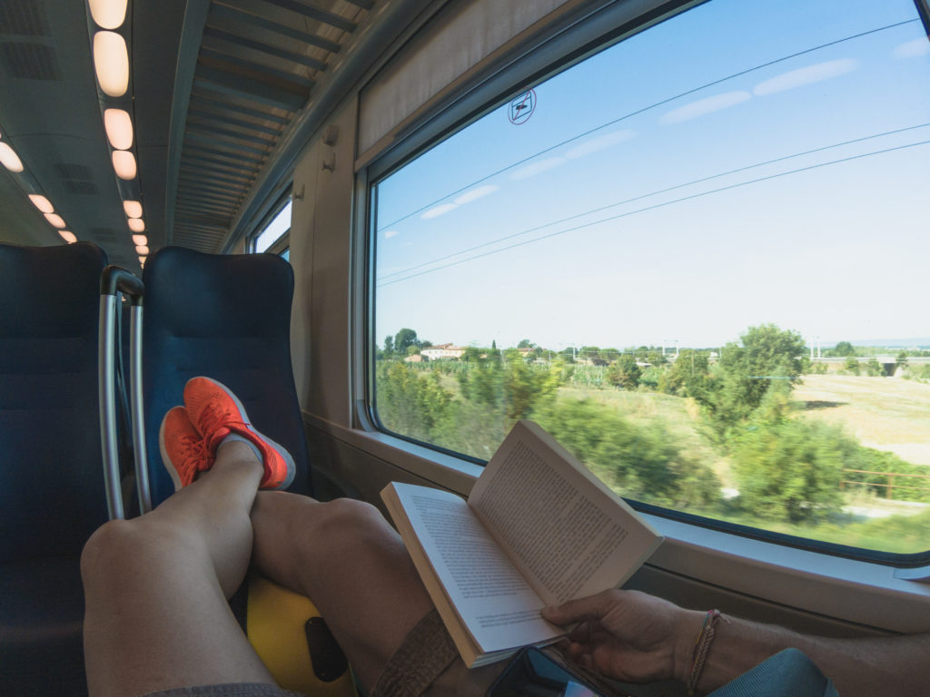 Vi ser beina til en person som sitter og ser ut av vinduet på et tog, med en bok i fanget.