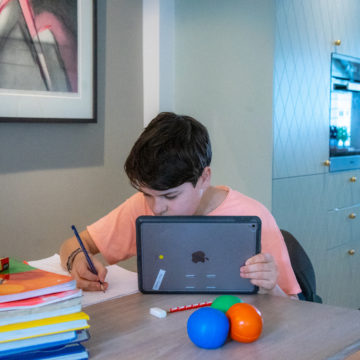 Gutt i barneskolealder gjør skoleoppgaver ved kjøkkenbordet. Han holder et nettbrett og skriver for hånd ved siden av. Skolebøker og noen fargerike leker på bordet.