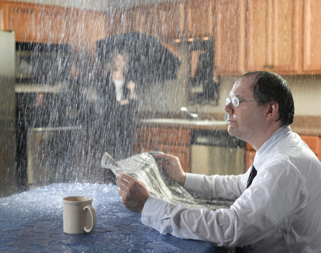 Regn faller ned på en mann som sitter på kjøkkenet sitt og leser avisa. I bakgrunnen: kvinne med paraply