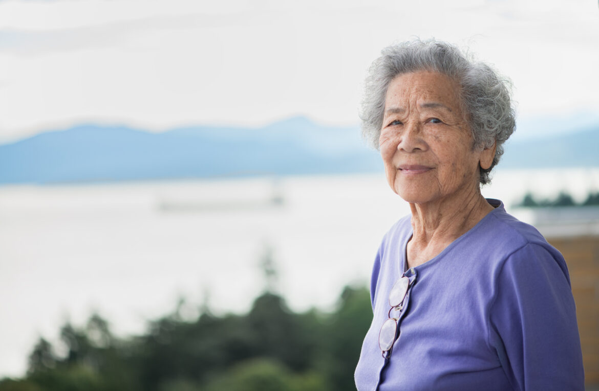 Eldre kvinne med grått hår og fiolett genser ser rett i kamera. I bakgrunnen ser vi en innsjø og fjell.