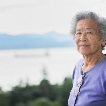 Eldre kvinne med grått hår og fiolett genser ser rett i kamera. I bakgrunnen ser vi en innsjø og fjell.